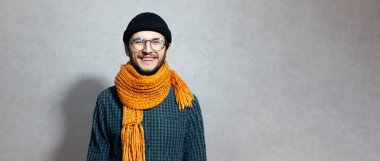 Portakal rengi eşarplı, gözlüklü, gri arka planda bereli ve gömlekli gülümseyen genç adam portresi..