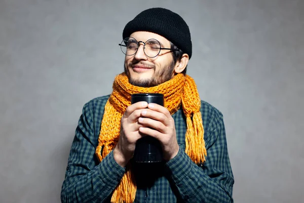Portrait of young man in green shirt with orange scarf and black hat, wearing glasses, warming hands with black coffee mug in hands.