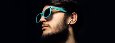 Cyan güneş gözlüğü ve kablosuz kulaklık takan erkek yüzünün yakın plan portresi. Siyah arkaplanda.