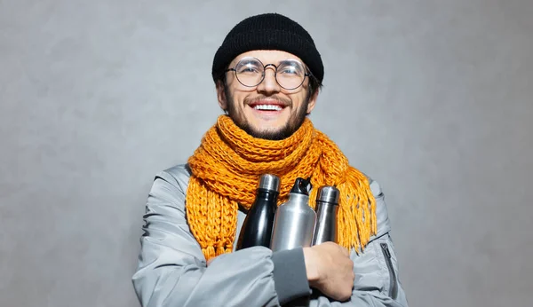 Portrait of young smiling man with knitted orange scarf, wearing glasses, black beanie hat and grey bomber, holding steel and aluminium thermo eco bottles for water.