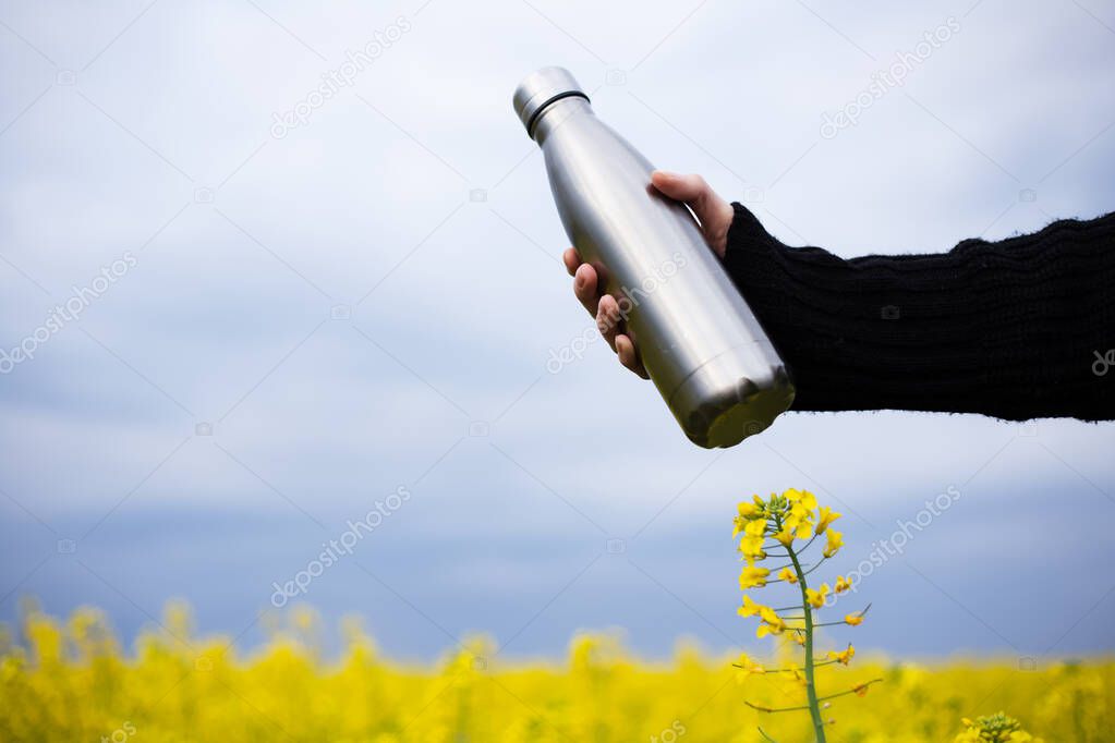 Stainless thermos bottle in hand, on background of rapeseed field
