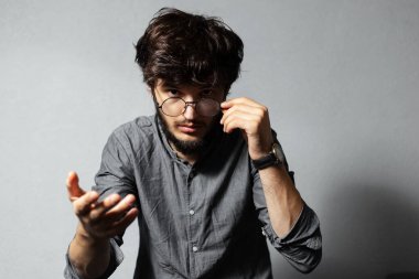 Dağınık saçlı genç sakallı adamın portresi, gözlüklerine dokunuyor, kameraya bakıyor ve işaret ediyor, gri arka planda..