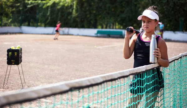 テニスコートの十代のスポーツ少女の肖像画 手に水とラケット用の再利用可能な熱ボトルでグリッドにもたれて — ストック写真