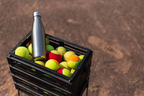 Black box with a lot of tennis balls and one steel thermo bottle for water. On tennis court. Say no to plastic bottles.