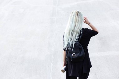 Sırt çantalı, siyah giyinmiş, elinde çelik termosu olan genç sarışın kız. Açık gri duvarın arka planında.