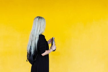 Plastiğe hayır de! Sarı arka planda siyah sırt çantalı, çelik şişe tutan sarışın genç bir kız..