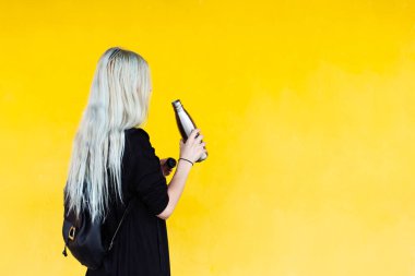 Plastiğe hayır de! Sarı arka planda siyah sırt çantalı, çelik şişe tutan sarışın genç bir kız..