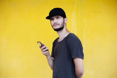 Sarı arka planda elinde akıllı telefon olan genç adamın portresi. Siyah tişört ve şapka giyiyor..