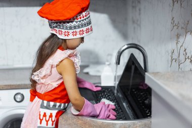 Çocuk lavaboda dizüstü bilgisayar yıkıyor. Kırmızı önlük, aşçı şapkası ve pembe eldivenler giyiyor..