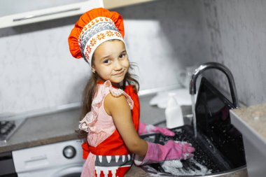 Çocuk lavaboda dizüstü bilgisayar yıkıyor. Kırmızı önlük, aşçı şapkası ve pembe eldivenler giyiyor..