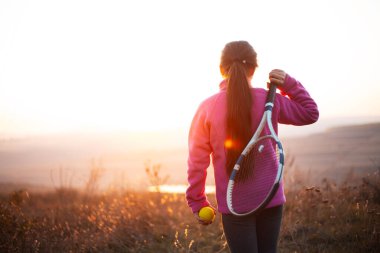 Küçük kızın arkası, tenis kortunun dışında gün batımında sahada antrenman becerisi. Pembe kazak giyip, tenis raketini top ile tutuyordu..