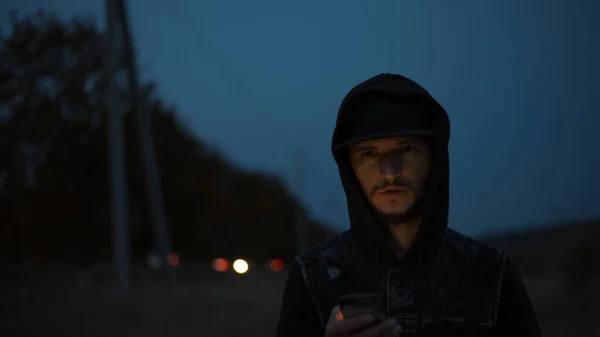 Dark portrait of young serious guy who stands on the street at night with smartphone in hands, wearing black cap and hooded sweater.