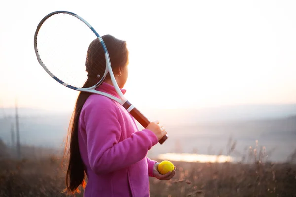 小女孩的画像是在日落时在网球场外的田里训练技巧 穿着粉色毛衣 拿着球拍 — 图库照片