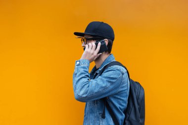 Portakal rengi arka planda akıllı telefondan konuşan genç hipster portresi. Kot ceket, kep, sırt çantası ve gözlük giyiyor..