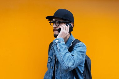 Portakal rengi arka planda akıllı telefondan konuşan genç hipster portresi. Kot ceket, kep, sırt çantası ve gözlük giyiyor..