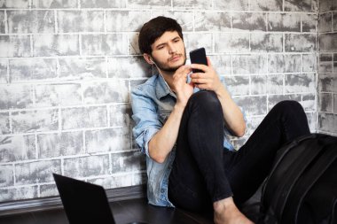 Genç esmer adam dizüstü bilgisayarı ve gri tuğla duvarın arkasında akıllı telefonuyla yerde oturuyor..