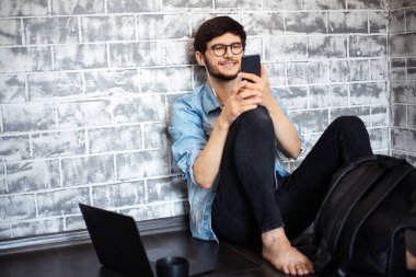 Genç esmer adam dizüstü bilgisayarı ve gri tuğla duvarın arkasında akıllı telefonuyla yerde oturuyor..