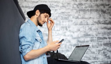 Kot ceketli ve siyah şapkalı bir gencin portresi yerde oturuyor, dizüstü bilgisayarda çalışıyor, akıllı telefon kullanıyor, siyah kahve içiyor..