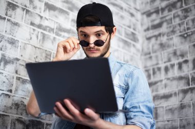 Gri tuğla duvarda siyah kapaklı ve güneş gözlüklü bir dizüstü bilgisayara bakan genç adamın portresi..
