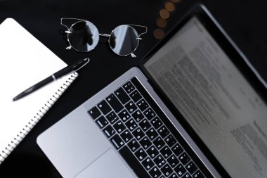 Dizüstü bilgisayarı, defteri, kalemi ve aynalı güneş gözlüğü olan modern, siyah çalışma masasının üst görüntüsü.