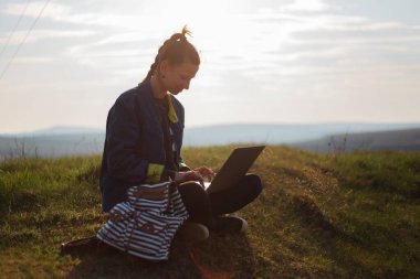 Kot ceketli genç hippi kız portresi, sahada günbatımının arka planında sırt çantasına kulaklık takan dizüstü bilgisayar kullanıyor..