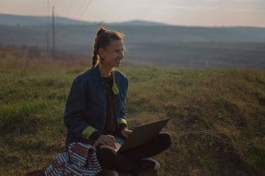 Kot ceketli genç hippi kız portresi, sahada günbatımının arka planında sırt çantasına kulaklık takan dizüstü bilgisayar kullanıyor..