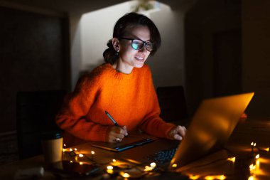 Dizüstü bilgisayarla grafik tablette çalışan gülümseyen tasarımcı kız portresi. Gözlüklü ve turuncu kazaklı. Karanlık oda evinde, çelenklerle.