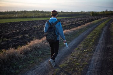 Sırt çantalı genç bir adam elinde alüminyum su şişesiyle köy yolunda yürüyor..
