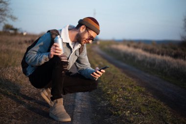 Sırt çantası, akıllı telefonu ve yeniden kullanılabilir alüminyum su şişesi olan hippi bir gencin portresi..