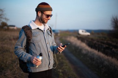 Sırt çantası, akıllı telefonu ve yeniden kullanılabilir alüminyum su şişesi olan hippi bir gencin portresi..