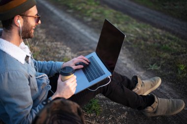 Dizüstü bilgisayarlı ve elinde bir fincan kahveyle oturan genç adamın yakın çekimi..