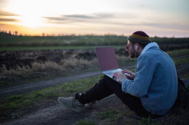Millennial hipster dizüstü bilgisayarda çalışan kulaklıklı adam gün batımında sahada..