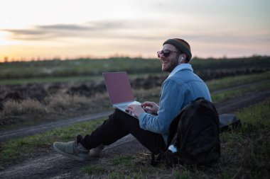 Millennial hipster dizüstü bilgisayarda çalışan kulaklıklı adam gün batımında sahada..