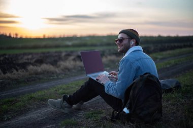 Millennial hipster dizüstü bilgisayarda çalışan kulaklıklı adam gün batımında sahada..