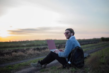 Millennial hipster dizüstü bilgisayarda çalışan kulaklıklı adam gün batımında sahada..