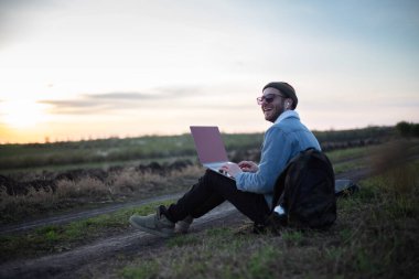 Millennial hipster dizüstü bilgisayarda çalışan kulaklıklı adam gün batımında sahada..