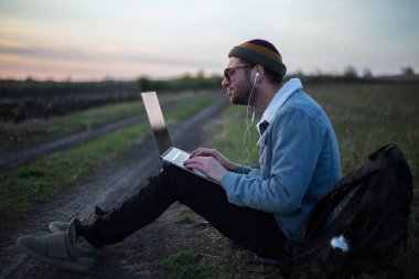 Millennial hipster dizüstü bilgisayarda çalışan kulaklıklı adam gün batımında sahada..