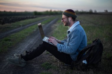 Millennial hipster dizüstü bilgisayarda çalışan kulaklıklı adam gün batımında sahada..