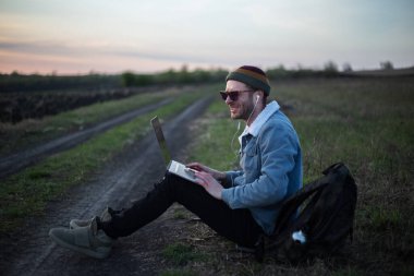 Millennial hipster dizüstü bilgisayarda çalışan kulaklıklı adam gün batımında sahada..