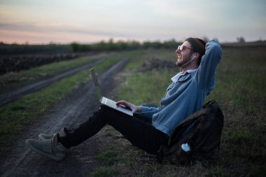Millennial hipster dizüstü bilgisayarda çalışan kulaklıklı adam gün batımında sahada..