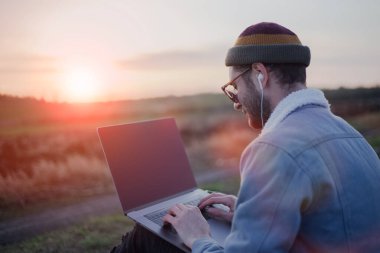Dizüstü bilgisayarı ve kulaklığı olan kendine güvenen hipster portresi, gün batımında çalışıyor, klavyede daktilo kullanıyor..