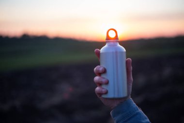 Gün batımının arka planında su için alüminyum şişeyi tutan bir adam..