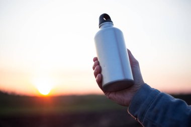 Gün batımının arka planında su için alüminyum şişeyi tutan bir adam..