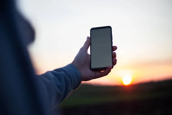 Mladý Muž Drží Smartphone Prázdným Bílým Mockup Pozadí Západu Slunce — Stock fotografie