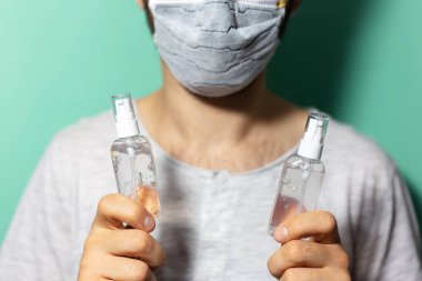 Erkek ellerinin iki şişe dezenfektan jeli tutuşu. Koronavirüse karşı antiseptik. Cyan, aqua menthe renginde tıbbi yüz maskesi takıyor..