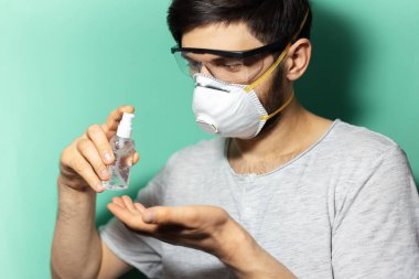 Delikanlının antiseptik jel önleme koronavirüsü kullanan, tıbbi maske takan ve arka planda su menilerinin renginde koruyucu gözlük takan yakın plan portresi..