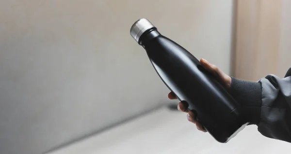 Ofis masasının arka planında kadın elinin yeniden kullanılabilir eko-çelik termodinamik su şişesini tutarken yakın plan..