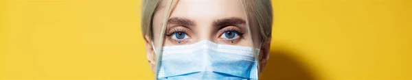 Panoramik portre, mavi gözlü genç kızın koronavirüse karşı güvenlik maskesi takması, sarı arka planda..
