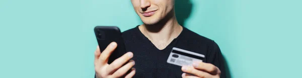 Panoramic close-up portrait of smiling guy holding smartphone and credit bank card on background of cyan, aqua menthe color.
