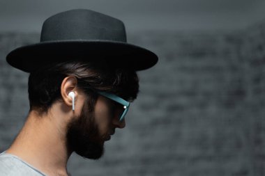 Siyah şapkalı, siyah güneş gözlüklü, gri arka planda kablosuz kulaklık kullanan genç adamın koyu renkli portresi..